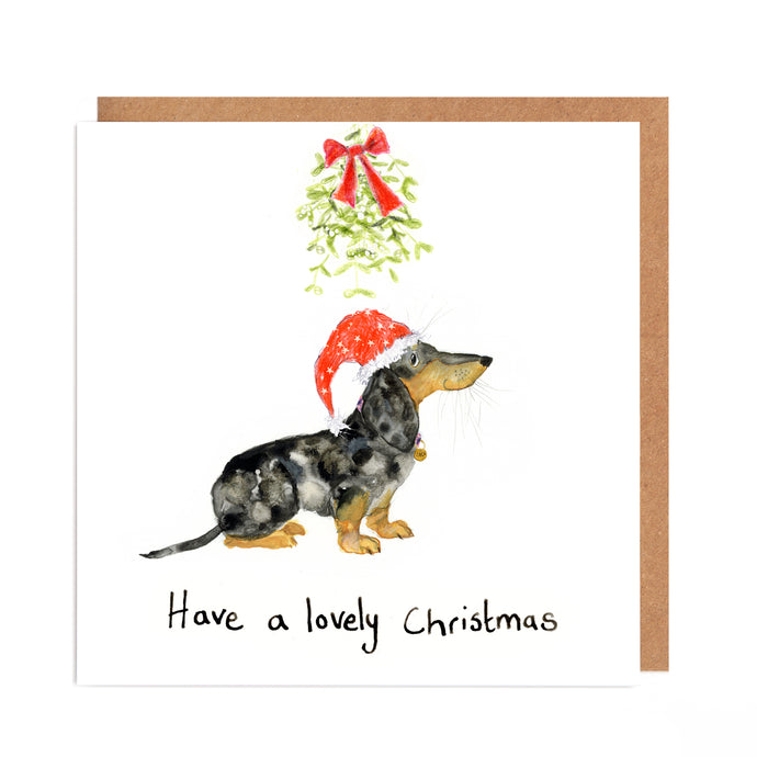 Square white dog Christmas card featuring watercolour and pencil illustration of a dachshund wearing a Christmas hat below some mistletoe tied with a red ribbon, with a handwritten 'have a lovely christmas' message below. 