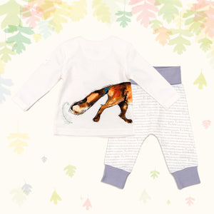 Baby gift set comprising a white top showing the tail of an illustrated wraparound fox print and matching bottoms with purple cuffs, on a backdrop of pastel leaves. 