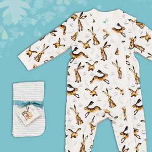 Baby gift set comprising a white babygrow featuring an illustrated hare and dragonfly print and matching dribble cloths tied in turquoise ribbon, on a blue backdrop with decorative  leaves. 