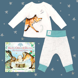 Baby gift set featuring a book from the Molly, Olive and Dexter series by Catherine Rayner, 'The Guessing Game' and a matching fox pyjama set laid on a midnight blue backdrop with silver stars. The pyjama set comprises a white long-sleeved top featuring an illustrated wraparound fox print and matching bottoms with blue cuffs.