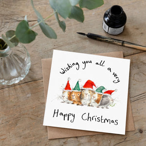 Christmas card featuring illustration of guinea pigs wearing red and green  christmas hats laid flat on a wooden table with a matching brown envelope. 