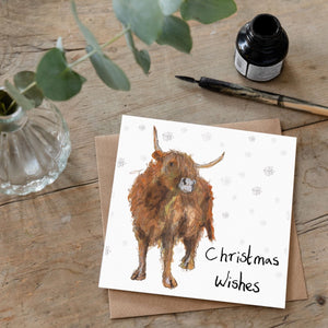 Christmas card laid flat on a wooden table with an illustration of Adrian the Highland Cow and handwritten message reading 'Christmas Wishes'