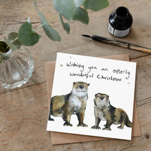 Load image into Gallery viewer, Christmas card featuring illustration of two otters and pun message &#39;wishing you an otterly wonderful christmas&#39; laid flat on a wooden table with a matching brown envelope. 