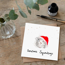 Load image into Gallery viewer, Christmas card featuring illustration of a guinea pig wearing a Santa hat laid flat on a wooden table with a matching brown envelope. 