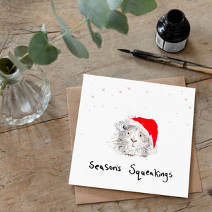 Christmas card featuring illustration of a guinea pig wearing a Santa hat laid flat on a wooden table with a matching brown envelope. 