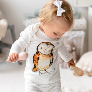 Blonde toddler standing up wearing Olive owl print long sleeve top and matching bottoms, looking down at wooden musical toys