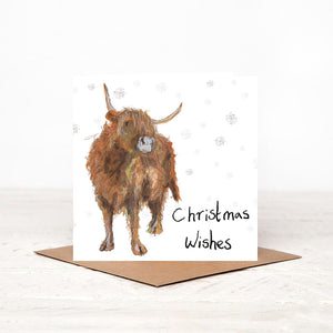 Square white Christmas card featuring Adrian the highland cow surrounding by snowflakes standing on top of the matching brown envelope. 